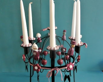 May include: A black metal candelabra with six candle holders, decorated with red and white fabric flowers and wooden figurines. The candelabra has a circular base and a central spire.