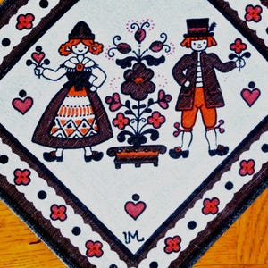Könnte beinhalten: Dekoratives quadratisches Textil mit einem Volkskunstdesign. Das Bild zeigt einen Mann und eine Frau in traditioneller Kleidung, die Blumen halten, mit einem Blumenschmuck. Der Rand hat ein sich wiederholendes Muster aus roten Blumen und schwarzen Punkten.