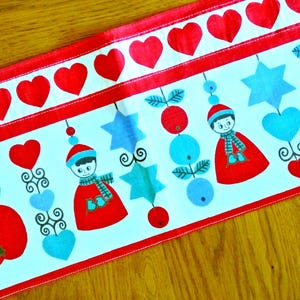 May include: A white fabric table runner featuring a repeating pattern of red hearts along the top border. The main section displays a colorful print of stylized figures in red hats and scarves, bright red apples, light blue stars, and hearts.