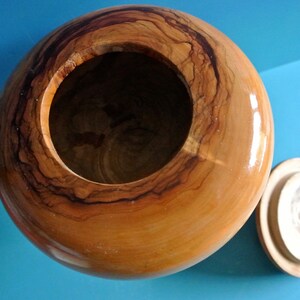 May include: A wooden bowl with a lid, both made of light brown wood with dark brown grain patterns. The bowl is round and has a smooth, polished finish.