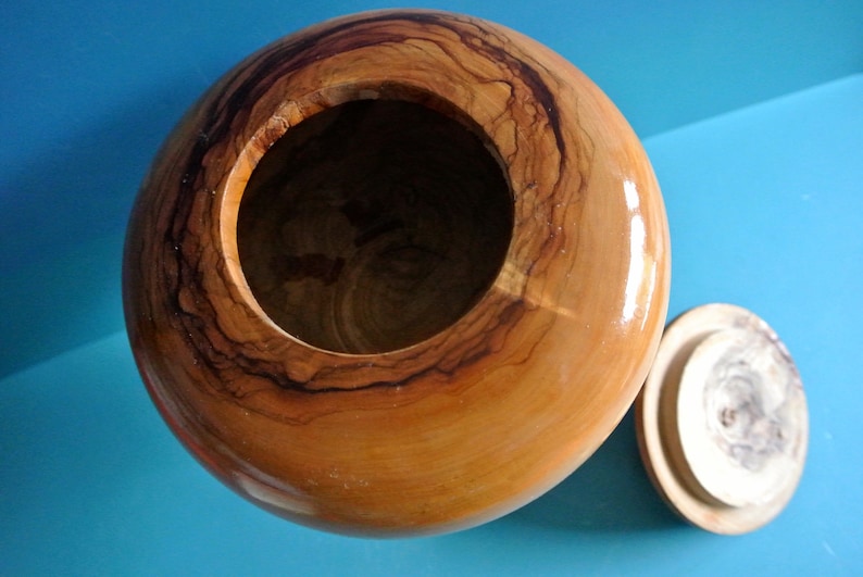 May include: A wooden bowl with a lid, both made of light brown wood with dark brown grain patterns. The bowl is round and has a smooth, polished finish.