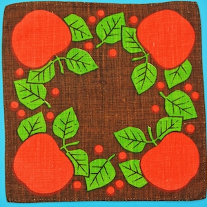 May include: A brown fabric square with a repeating pattern of red apples and green leaves. The apples are stylized and have a simple shape. The leaves are also stylized and have a simple shape. The fabric has a textured surface.