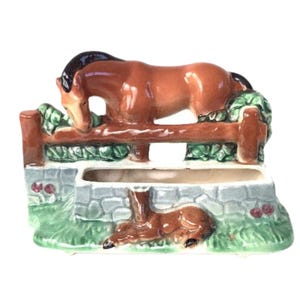 May include: A ceramic planter featuring a brown horse leaning over a wooden fence, with a brown foal lying below. The planter has a stone wall base with green grass and foliage accents. The horse has a black mane.