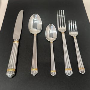 Puede incluir: Un juego de cubiertos plateados con detalles dorados. El conjunto incluye un cuchillo, dos cucharas y dos tenedores. Los mangos tienen un diseño acanalado y están adornados con bandas doradas. Los cubiertos están dispuestos sobre una superficie oscura.