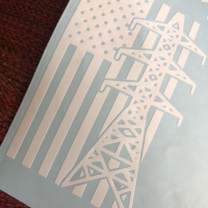 May include: White vinyl decal featuring an American flag design with a cutout star field and stripes. A stylized power line tower is superimposed on the flag. The decal is on a light blue background.