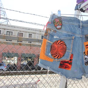 Puede incluir: Pantalones cortos de mezclilla azul con varios parches, incluyendo una calavera con flores, un diseño de espiral, una caja torácica y un diseño de llama. Los pantalones cortos tienen un aspecto desgastado y están colgados en un tendedero.