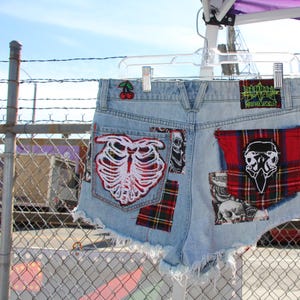 Puede incluir: Pantalones cortos de mezclilla azul claro con parches, incluyendo una caja torácica roja y blanca, una tela de cuadros rojos y negros y una calavera negra y blanca. Los pantalones cortos tienen un parche de cereza en el bolsillo trasero y el texto "Monster World" en la parte posterior.