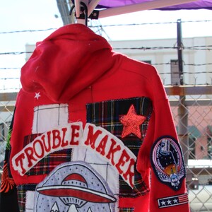 Puede incluir: Sudadera roja con capucha con un diseño de patchwork que incluye un OVNI, montañas, tela a cuadros y el texto "Trouble Maker".