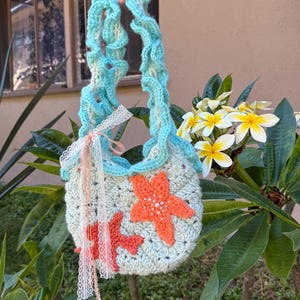May include: A handmade crochet bag with a light blue and white body, adorned with two orange starfish appliqués. The bag features a ruffled light blue handle and is embellished with lace and ribbons. The bag is held up by a hand.