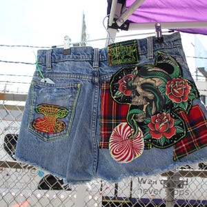 Puede incluir: Un par de pantalones cortos de mezclilla con varios parches cosidos. Los parches incluyen una calavera con una serpiente, un patrón de cuadros rojos y negros, un diseño de espiral rojo y blanco y un diseño geométrico. Los pantalones cortos están deshilachados en la parte inferior.
