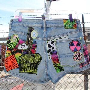 Puede incluir: Pantalones cortos de mezclilla azul claro con varios parches coloridos, que incluyen un diseño de hongo, una impresión de vaca y un símbolo rosa y negro. El texto "why not?" es visible en uno de los parches.