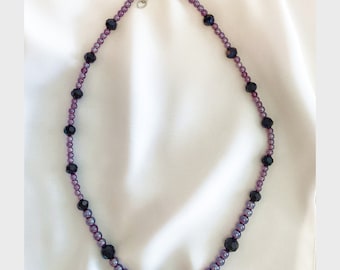 Puede incluir: Un collar de cuentas de color morado y negro con un cierre plateado. El collar está hecho de cuentas pequeñas y redondas.