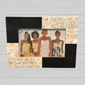 May include: A black and white picture frame made of mother of pearl and wood. The frame has a photo of four people in it. The photo shows four people in dresses, standing in a line.