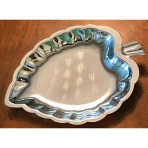 May include: A silver-coloured leaf-shaped serving tray with a scalloped edge. The tray has a smooth, reflective surface and a small stem detail. The tray is likely made of metal and is designed for serving food or displaying items.