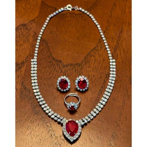 May include: A silver-coloured jewellery set comprising a necklace, earrings, and a ring. The necklace and earrings have a double-row design with clear stones. The ring and necklace pendant feature a large, red, teardrop-shaped stone.