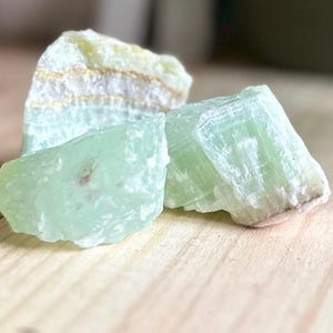May include: Three raw green calcite mineral specimens. The calcite pieces have a crystalline structure and varying shades of green, with some areas showing white and gold striations. The stones are displayed on a light wooden surface.