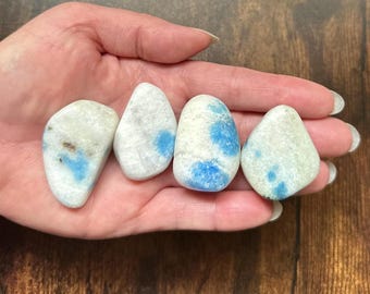 May include: Four tumbled stones with a mix of white and blue coloring are held in a hand. The stones have a smooth, polished appearance and vary slightly in shape. The blue coloration appears in patches and spots across the stones.