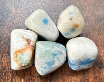 May include: Five tumbled stones in shades of white, blue, and brown. The stones have a smooth, polished appearance with unique patterns and markings. The stones are arranged on a wooden surface.