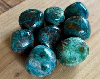 May include: A collection of polished, tumbled stones in shades of teal, turquoise, and brown. The stones have a smooth, glossy finish and are arranged on a light-colored wooden surface. The stones vary in size and shape, with a mix of colors and patterns.