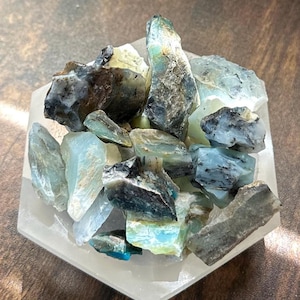 May include: A collection of raw, uncut gemstones in shades of blue, green, and gray, resting on a white, hexagonal dish. The stones vary in size and shape, showcasing natural textures and colors. The arrangement creates a visually appealing display.