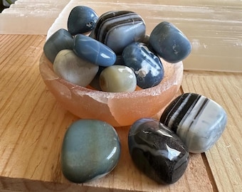 May include: A collection of polished gemstones in shades of blue, gray, and black, displayed in a pink bowl. The stones vary in size and pattern, with some featuring stripes and others solid colors. The bowl sits on a wooden surface, with a white rectangular object in the background.
