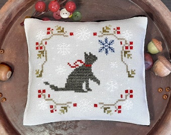 May include: A white cross-stitch cushion featuring a black cat design, a red ribbon, and a snowflake. The cushion is surrounded by a border of red berries and green leaves. The background is a light brown wooden surface.