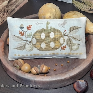 Puede incluir: Una almohada blanca con un diseño bordado de una calabaza con hojas y bellotas. La almohada está sentada en un tazón de madera marrón con otras calabazas y bellotas.