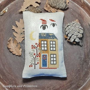 May include: A hand-stitched pillow featuring a whimsical house with a blue door, a crescent moon, and two black cats wearing red hats. The house is yellow with blue and white trim and has three windows. The pillow is on a brown surface with pine cones and leaves.