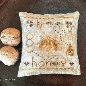 May include: A beige pillow with a cross-stitch design featuring a bee, honeycomb, and the word "Honey".