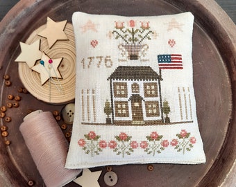 May include: A cream-colored, cross-stitched pillow featuring a colonial house with an American flag. The design includes the year "1776", floral accents, and a vase of flowers. Wooden stars, beads, and thread are arranged around the pillow.