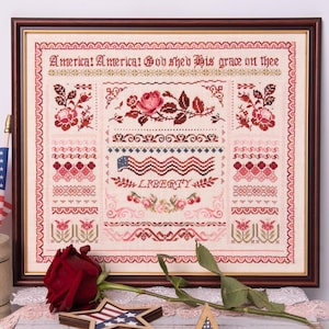 Può includere: Campione a punto croce incorniciato a tema patriottico. Il disegno presenta il testo "America! America! God shed His grace on thee", una bandiera americana e la parola "LIBERTY". Il campione è nei toni del rosso, rosa e crema.