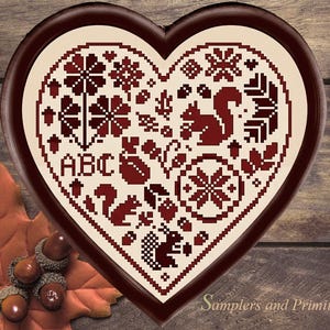May include: A heart-shaped cross-stitch sampler featuring dark red thread on a cream background. The design includes squirrels, acorns, leaves, a snowflake-like pattern, and the letters ABC. The sampler is enclosed in a dark brown wooden frame.