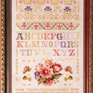 Op de afbeelding: Een ingelijste kruissteek sampler met bloemen- en geometrische patronen. Het ontwerp bevat een bloemenboeket, alfabetletters en decoratieve randen in roze, paars, groen en goud. De lijst heeft een sierlijke gouden afwerking.