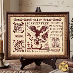 May include: A framed cross-stitch sampler with a patriotic theme. The design features an eagle, American flag, and the text "Forever Free" and "Liberty and Union." The color palette is primarily burgundy and cream, with decorative borders and floral elements.