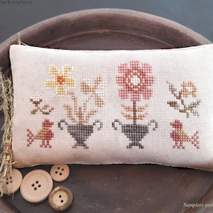Peut inclure: Un coussin blanc avec un motif de point de croix représentant trois fleurs en pot et deux oiseaux. Le coussin est posé sur une assiette en terre cuite brune avec des boutons.