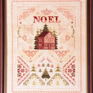 Op de afbeelding: Ingeklemd kruissteekkunstwerk met een huis en bomen, met het woord "NOEL" in het rood. Het ontwerp bevat geometrische patronen en bloemelementen in tinten rood, groen en beige. Het kunstwerk is ingelijst in een donkerbruine houten lijst.
