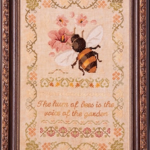 May include: Cross-stitch picture in a dark brown frame, depicting a bee with orange and black stripes near pink flowers. The text reads "The hum of bees is the voice of the garden". The frame has an ornate, decorative design.