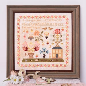 Puede incluir: Un sampler de punto de cruz enmarcado con un tema de jardín. El diseño incluye flores, pájaros, una casa y el texto "In the garden", junto con el alfabeto y números. El marco es de color marrón oscuro.