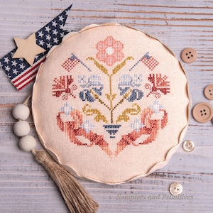 Puede incluir: Un cojín redondo de punto de cruz color crema con un diseño floral en rojo, azul y dorado. Dos pequeñas banderas americanas están cosidas en la parte superior. Una estrella de madera y una bandera americana de tela están a la izquierda.