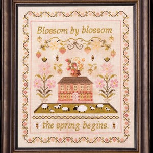 May include: Framed cross-stitch artwork featuring a house, sheep, and floral elements. The text "Blossom by blossom" and "the spring begins" are stitched in gold thread. The piece is framed in a dark brown wooden frame.