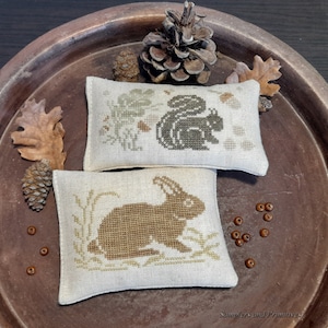 May include: Two embroidered pillow covers with a woodland theme. One features a brown squirrel with a leafy background and the other has a brown rabbit with green foliage. Both pillows are on a brown wooden surface.