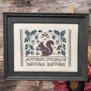 May include: A framed cross-stitch embroidery on off-white fabric featuring a brown squirrel holding an acorn, surrounded by dark green oak leaves and small acorns. The text "AUTUMN STORAGE" is stitched in brown below the squirrel design.