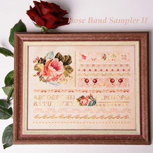 Pode incluir: Um sampler de ponto cruz emoldurado intitulado "Rose Band Sampler II" com padrões florais e geométricos em tons de rosa, verde e amarelo. O sampler inclui um alfabeto e motivos de rosas. Uma rosa vermelha é colocada acima da moldura.