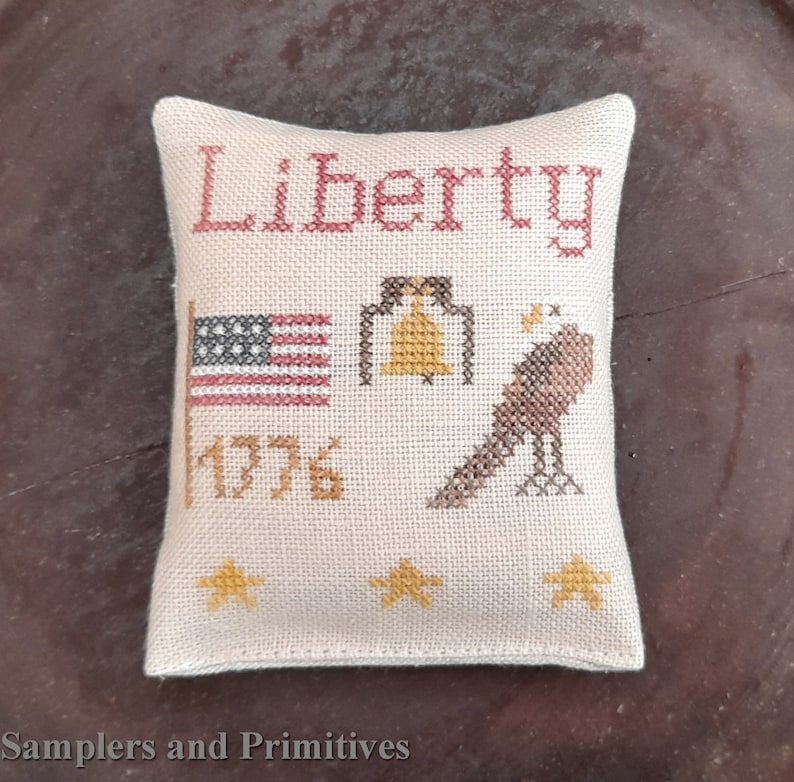 May include: A beige pillow with a cross-stitch design featuring the word "Liberty", an American flag, a bell, a bird, and the year "1776".