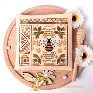 Puede incluir: Un sampler de punto de cruz con una abeja rodeada de panales y flores. El sampler está sobre una base redonda de madera con un cucharón de miel y flores.