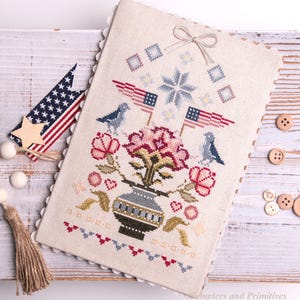 Puede incluir: Un sampler de punto de cruz con imágenes patrióticas. El diseño incluye banderas americanas, pájaros y flores en un jarrón. La paleta de colores es roja, blanca, azul y beige. También hay una estrella de madera y una borla.