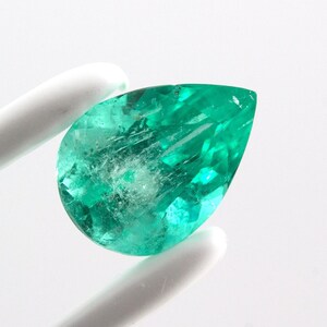 Puede incluir: Una piedra preciosa esmeralda de forma de pera con un tono verde, sostenida por pinzas sobre un fondo blanco.