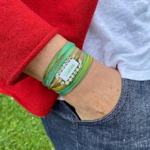 May include: A green, gold, and white bracelet with a white tag that says "Listen" and rhinestones. The bracelet is worn on a person's wrist.