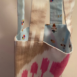 Könnte beinhalten: Eine Stoff-Tragetasche mit einem braunen und rosa Ikat-Muster auf der Außenseite. Das Innenfutter ist hellblau mit einem Strandmotiv. Die Tasche hat braune Bandgriffe.