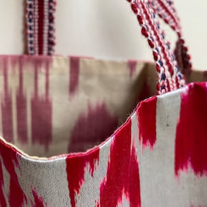 Könnte beinhalten: Eine rote und weiße Ikat-Stofftasche mit gewebten roten, weißen und blauen Henkeln. Die Tasche ist aus einem weichen, strukturierten Stoff.